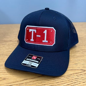 May include: A navy blue trucker cap featuring a rectangular red patch with white stitched borders and the white text T-1 in the centre. The cap includes a black sticker on the brim with the text Richardson Original 112 Trucker X-Large.