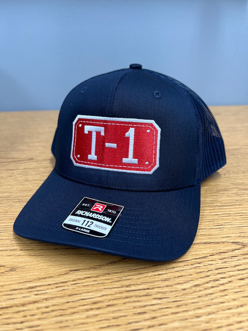 May include: A navy blue trucker cap featuring a rectangular red patch with white stitched borders and the white text T-1 in the centre. The cap includes a black sticker on the brim with the text Richardson Original 112 Trucker X-Large.