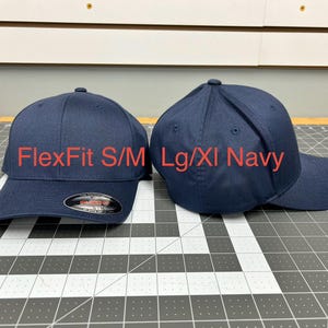 May include: Two navy blue baseball caps with a structured crown and curved brim. The left cap features a black and red sticker on the brim. Text on the image reads FlexFit S/M and Lg/Xl Navy, indicating the two available size options for these hats.