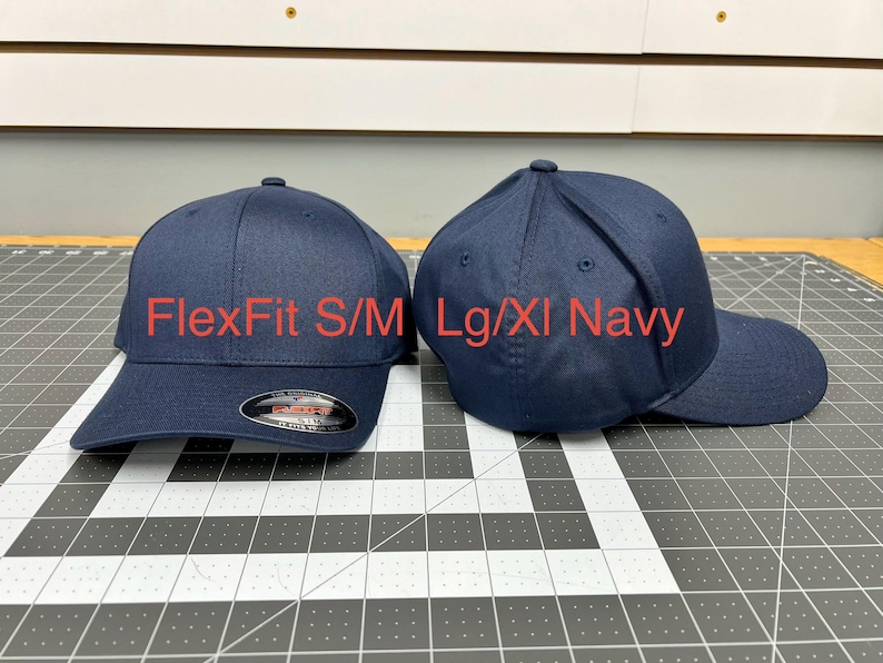 May include: Two navy blue baseball caps with a structured crown and curved brim. The left cap features a black and red sticker on the brim. Text on the image reads FlexFit S/M and Lg/Xl Navy, indicating the two available size options for these hats.