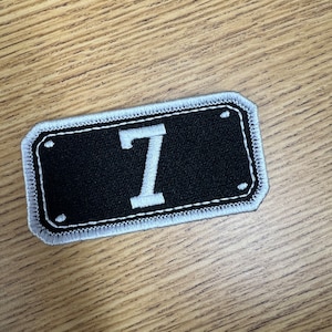 May include: A rectangular black patch with a white embroidered border. The number 7 is prominently displayed in white in the center. The patch has a stitched edge and is set against a wooden background.
