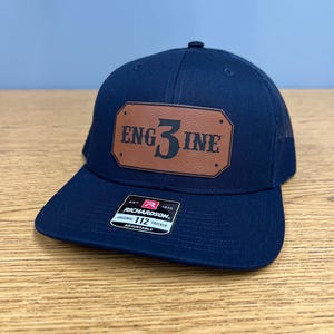 Peut inclure: Casquette de camionneur bleu marine avec un patch en cuir marron. Le patch porte l'inscription "ENG3INE". La casquette a une visière incurvée et un dos en filet. Elle est réglable et porte une étiquette Richardson.