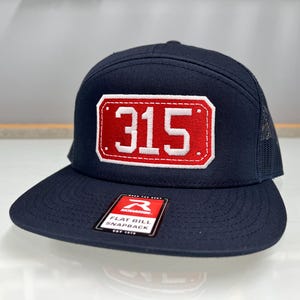 Könnte beinhalten: Marineblaue Flat-Bill-Snapback-Kappe mit einem rot-weißen, gestickten Aufnäher mit der Nummer "315". Die Kappe hat eine Mesh-Rückseite und einen flachen Schirm. Ein kleines Markenetikett ist sichtbar.