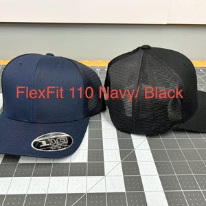 May include: Two baseball caps, one navy blue and one black, both featuring a mesh back panel and a structured crown. The navy cap displays a circular sticker on the brim with the text FlexFit 110. The text FlexFit 110 Navy/ Black is visible.