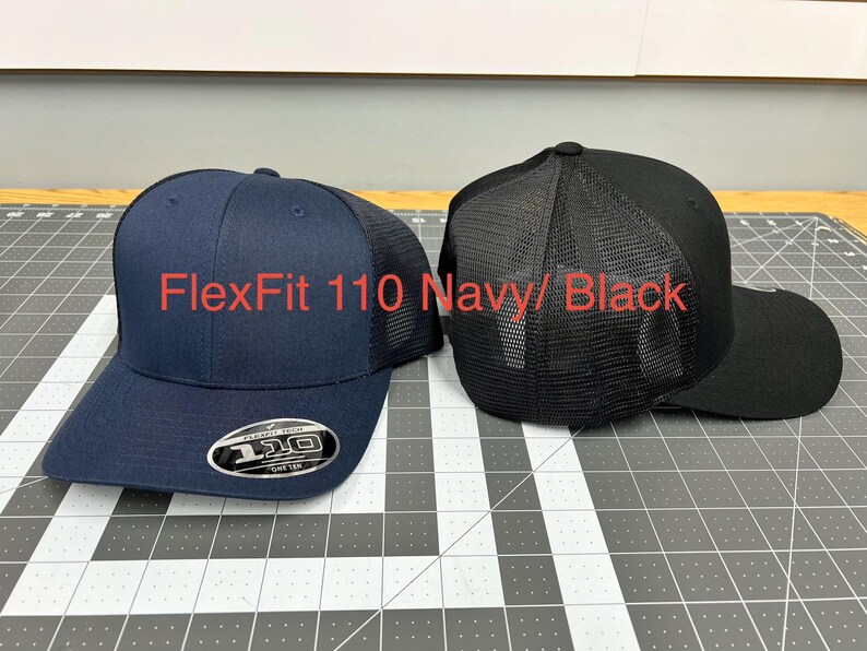 May include: Two baseball caps, one navy blue and one black, both featuring a mesh back panel and a structured crown. The navy cap displays a circular sticker on the brim with the text FlexFit 110. The text FlexFit 110 Navy/ Black is visible.