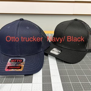 May include: Two Otto trucker hats, one navy blue and one black, featuring mesh back panels and solid front panels. Each hat has a sticker on the brim with the text Otto Five Star Trucker. The text Otto trucker Navy/ Black is visible.