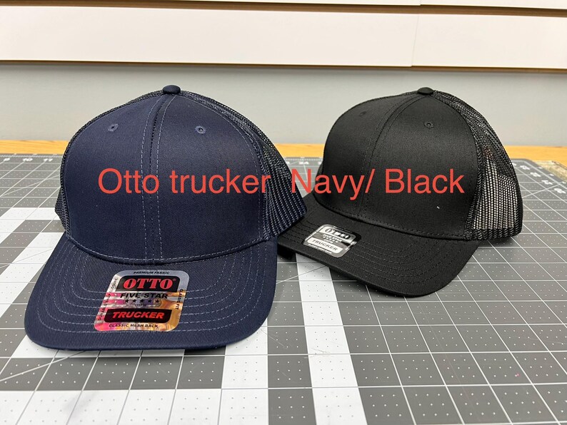 May include: Two Otto trucker hats, one navy blue and one black, featuring mesh back panels and solid front panels. Each hat has a sticker on the brim with the text Otto Five Star Trucker. The text Otto trucker Navy/ Black is visible.