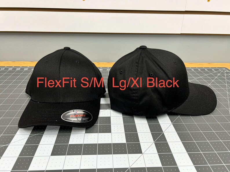 May include: Two solid black baseball caps with a structured crown and curved brim. The left cap features a silver and black sticker on the brim with the text FlexFit S/M. Red text overlay reads FlexFit S/M and Lg/Xl Black above the respective caps.