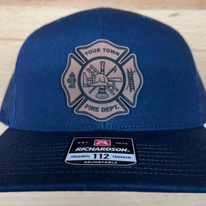 May include: A navy blue trucker hat with a brown leather patch featuring a firefighter's emblem. The patch has the text "Your Town Fire Dept." and depicts a firefighter's helmet, axe, ladder, hose, and fire hydrant. The hat has a black mesh back and a white adjustable strap with the text "Richardson 112 Original Trucker Adjustable" and the company logo.