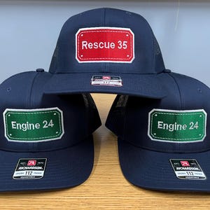 May include: Three navy blue trucker caps with mesh backs and flat peaks. The top cap features a red rectangular patch with white text reading Rescue 35. The two bottom caps feature green rectangular patches with white text reading Engine 24.