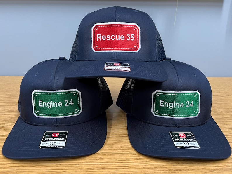 May include: Three navy blue trucker caps with mesh backs and flat peaks. The top cap features a red rectangular patch with white text reading Rescue 35. The two bottom caps feature green rectangular patches with white text reading Engine 24.