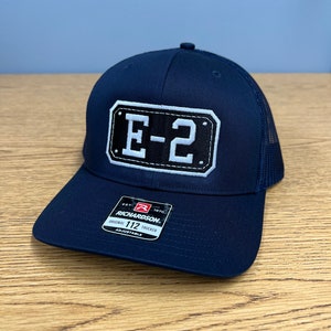 May include: A navy blue trucker hat featuring a rectangular patch on the front with the text E-2 in white letters on a black background. The hat includes a sticker on the brim with the text Richardson 112 Original Trucker Adjustable.