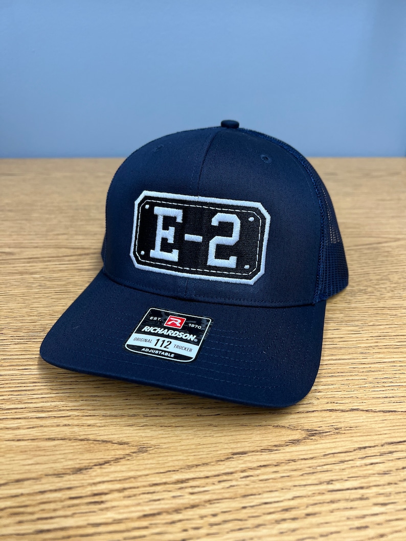 May include: A navy blue trucker hat featuring a rectangular patch on the front with the text E-2 in white letters on a black background. The hat includes a sticker on the brim with the text Richardson 112 Original Trucker Adjustable.