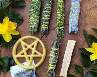 May include: A collection of spiritual items displayed on a wooden surface. Features bundles of dried herbs bound with yellow string, a yellow pentacle, a candle, a crystal, a bag of herbs, and a piece of palo santo. Yellow daffodils and green leaves are also present.