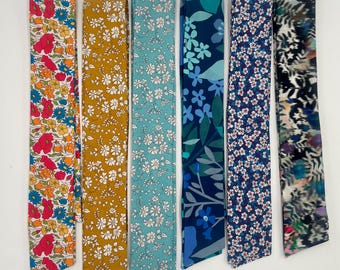 May include: Seven colorful fabric neck scarves are displayed. The scarves feature floral and abstract patterns in various colors, including red, yellow, blue, and teal. The scarves are rectangular and appear to be made of a lightweight material.