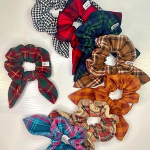 May include: A collection of scrunchies in various plaid patterns, including red, green, blue, and orange. One scrunchie features a black and white houndstooth design. Each scrunchie has a fabric bow and a small white tag.