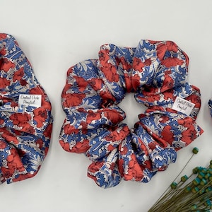 Könnte beinhalten: Drei Haargummis in verschiedenen Größen mit einem roten und blauen Blumenmuster. Jedes Haargummi hat ein weißes Etikett mit schwarzem Text. Die Haargummis sind aus glänzendem Material und auf einer weißen Oberfläche angeordnet.