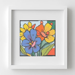 May include: A framed floral cross-stitch artwork. The artwork features a vibrant bouquet of flowers in shades of blue, yellow, and orange, set against a light blue and green background. The frame is white, and the artwork is displayed on a white wall.