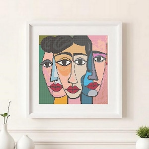 May include: Framed abstract art featuring three stylized faces with bold lines and vibrant colors. Each face has a unique color palette, including pink, blue, and yellow, with red lips. The artwork is in a white frame.