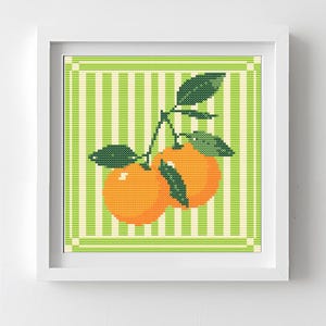 May include: A framed cross-stitch artwork featuring two oranges with green leaves on a green and white striped background. The artwork is in a white frame and is displayed against a white wall.