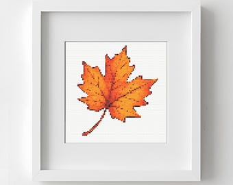 May include: A framed cross-stitch of a maple leaf in shades of orange and yellow. The leaf is centered on a white background within a white frame, evoking autumn. The artwork is displayed on a white wall.