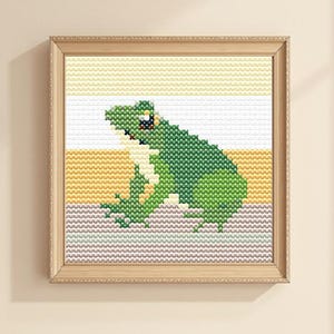 Könnte beinhalten: Ein gerahmtes Kreuzstich-Kunstwerk mit einem grünen Frosch. Der Frosch ist in Grüntönen dargestellt, mit roten Akzenten für die Augen, vor einem gestreiften Hintergrund aus Gelb, Weiß und Grau. Der Rahmen ist hellbraun.