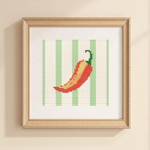 May include: A framed cross-stitch artwork depicting a red and yellow chilli pepper with a green stem. The pepper is set against a background of green and white vertical stripes. The frame is light wood.