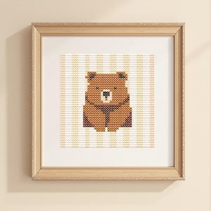 May include: A framed cross-stitch artwork featuring a brown bear. The bear is centered on a striped background of white and light yellow. The artwork is in a light brown wooden frame.