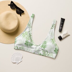 Op de afbeelding: Witte bikinitop met een groen palmbladpatroon. Onderdeel van een zomerse strandkledingcollectie, samen met een strohoed, zonnebril, zonnebrandcrème en een zeeschelp.