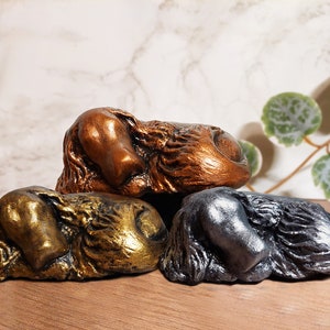 Peut inclure: Trois sculptures décoratives aux finitions métalliques : bronze, or et argent. Chaque sculpture représente un animal endormi stylisé, peut-être un chien ou une créature similaire. Les sculptures sont exposées sur une surface en bois.