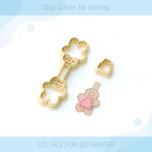 May include: A set of three 3D printed clay cutters for making earrings. The cutters are shaped like a flower, a dress, and a person. The cutters are made of a light brown plastic and are designed to be used with polymer clay.