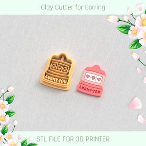 Puede incluir: Dos cortadores de arcilla para pendientes. Uno es de color canela, el otro rosa. Ambos tienen forma de máquina tragamonedas con tres símbolos de corazón y el texto "LUCKY YOU". El texto superior dice "Clay Cutter for Earring", y el texto inferior dice "STL FILE FOR 3D PRINTER".