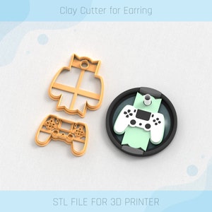 Puede incluir: Un cortador de arcilla impreso en 3D para pendientes, con forma de mando de videojuego. El cortador es naranja y tiene un agujero para colgarlo. La imagen también muestra una base redonda negra con un mando verde y blanco en la parte superior. El texto "STL FILE FOR 3D PRINTER" está en la parte inferior de la imagen.
