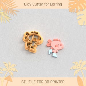 Puede incluir: Un cortador de arcilla y un pendiente terminado con forma de pez de dibujos animados. El cortador es de color naranja claro y el pendiente es rosa con detalles azules. El texto en la parte superior dice "Clay Cutter for Earring" y en la parte inferior "STL FILE FOR 3D PRINTER".