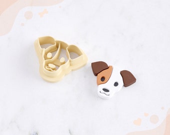 May include: A 3D printed dog head clay cutter for earrings. The cutter is beige and the printed dog head is white with brown ears and a brown patch on its face. The text "STL FILE FOR 3D PRINTER" is at the bottom of the image.