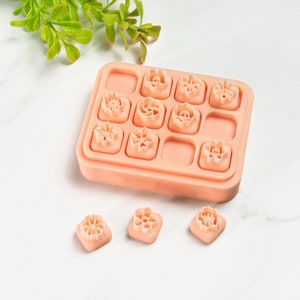 Pode incluir: Um conjunto de 10 cortadores de biscoitos de silicone rosa em forma de flor em uma bandeja retangular. Três cortadores adicionais são mostrados separadamente em uma superfície branca.