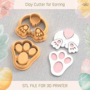 May include: A set of four 3D printed clay cutters for making bunny-shaped earrings. The cutters are in the shape of a bunny's back, a bunny's paw, and two bunny ears. The cutters are made of white plastic and have a light pink colour on the inside. The text "STL FILE FOR 3D PRINTER" is printed on a brown surface below the cutters.