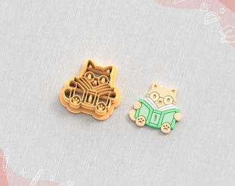 Könnte beinhalten: Ein hellbrauner Ton-Ausstecher und ein fertiger Ohrring, beide in Form einer Katze, die ein Buch liest und eine Brille trägt. Der Ausstecher ist hellbraun und der Ohrring ist hellbraun, grün und weiß. Der Text "Clay Cutter for Earring" und "STL FILE FOR 3D PRINTER" sind sichtbar.