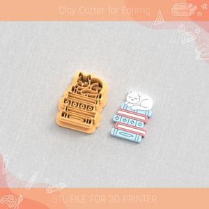 Puede incluir: Cortador de arcilla para pendientes con forma de gato durmiendo sobre una pila de libros. El cortador es de color naranja claro, y el pendiente terminado es blanco, azul y rosa. El texto "Clay Cutter for Earring" y "STL FILE FOR 3D PRINTER" son visibles.