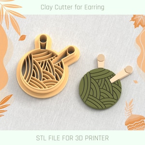 May include: A 3D printed clay cutter for earrings, shaped like a ball of yarn with two knitting needles. The cutter is beige and the yarn is green.