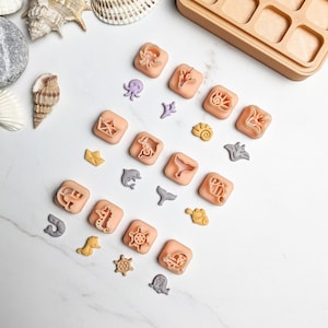 Op de afbeelding: Een set van 18 plastic koekjesvormen in verschillende vormen met zeethema, waaronder een walvis, een dolfijn, een zeester, een schelp, een krab, een octopus, een zeilboot, een zeepaardje en een stuurwiel. De vormen zijn gerangschikt op een wit oppervlak.