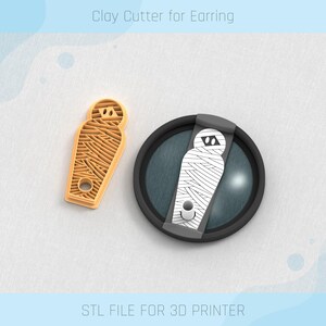 May include: A 3D printed clay cutter for earrings shaped like a mummy. The cutter is made of a light brown plastic and has a hole in the center. The cutter is shown next to a black and white 3D printed model of a mummy.