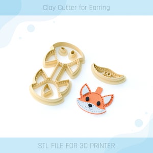 Könnte beinhalten: Ein 3D-gedruckter Ton-Ausstecher für Ohrringe mit einem Fuchs-Design. Der Ausstecher besteht aus beigefarbenem Kunststoff und enthält ein separates Stück für die Fuchsnase. Das Gesicht des Fuchses ist orange mit schwarzen Augen und einer weißen Schnauze. Der Text "STL FILE FOR 3D PRINTER" befindet sich am unteren Rand des Bildes.