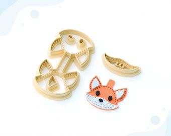 May include: A 3D printed clay cutter for earrings, featuring a fox design. The cutter is made of beige plastic and includes a separate piece for the fox's nose. The fox's face is orange with black eyes and a white muzzle. The text "STL FILE FOR 3D PRINTER" is at the bottom of the image.