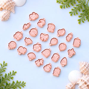 May include: A collection of small, light pink cookie cutters in various shapes, including flowers, shells, and fruit, arranged on a light blue surface. The cutters are made of plastic and are ideal for baking. Seashells and greenery are in the background.