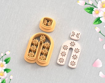 Pode incluir: Dois cortadores de argila impressos em 3D para brincos, um em forma de U com um design floral e o outro em forma de quadrado pequeno. Os cortadores estão sobre um fundo de tecido marrom claro. ARQUIVO STL PARA IMPRESSORA 3D.