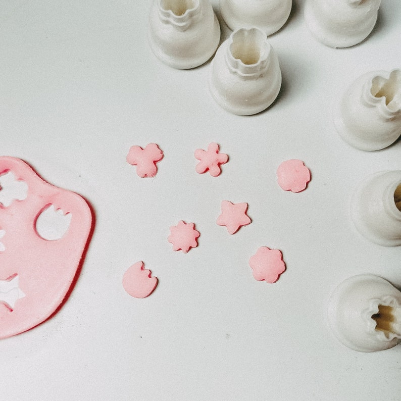 Może przedstawiać: R&oacute;żowe wykrojniki z fondantu w r&oacute;żnych kształtach, w tym gwiazdki, kwiaty i tulipan, na białym tle. Białe plastikowe foremki do ciasteczek w tle.