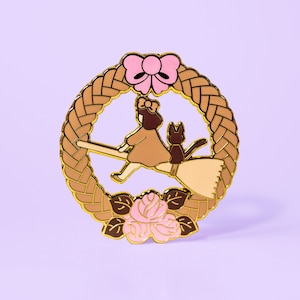May include: Gold-tone enamel pin featuring a girl and a cat flying on a broomstick, surrounded by a brown braided wreath with pink roses and a pink bow.