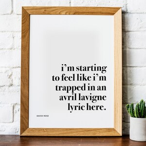 May include: A framed print with a white background and black text that reads "I'm starting to feel like I'm trapped in an Avril Lavigne lyric here." The frame is made of light brown wood. The author is David Rose.