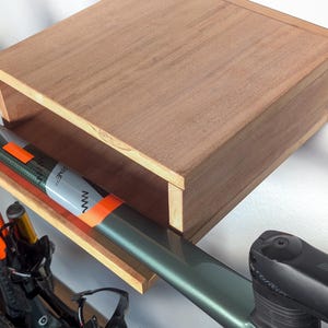 Peut inclure: Une étagère en bois avec un vélo suspendu. L'étagère est fixée à un mur blanc. Le vélo a un cadre noir avec des accents orange et vert. Le vélo a un guidon noir et une selle noire.
